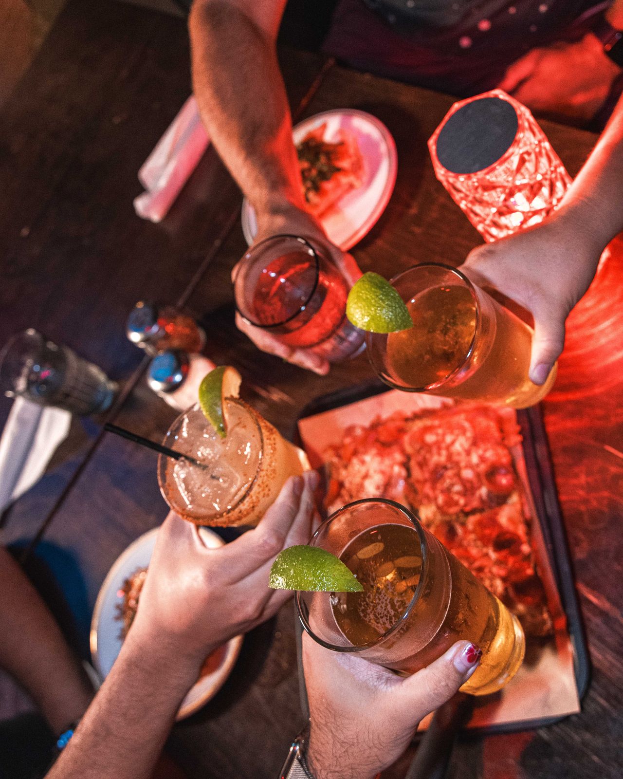 An overhead shot of four hands toasting drinks over a pizza