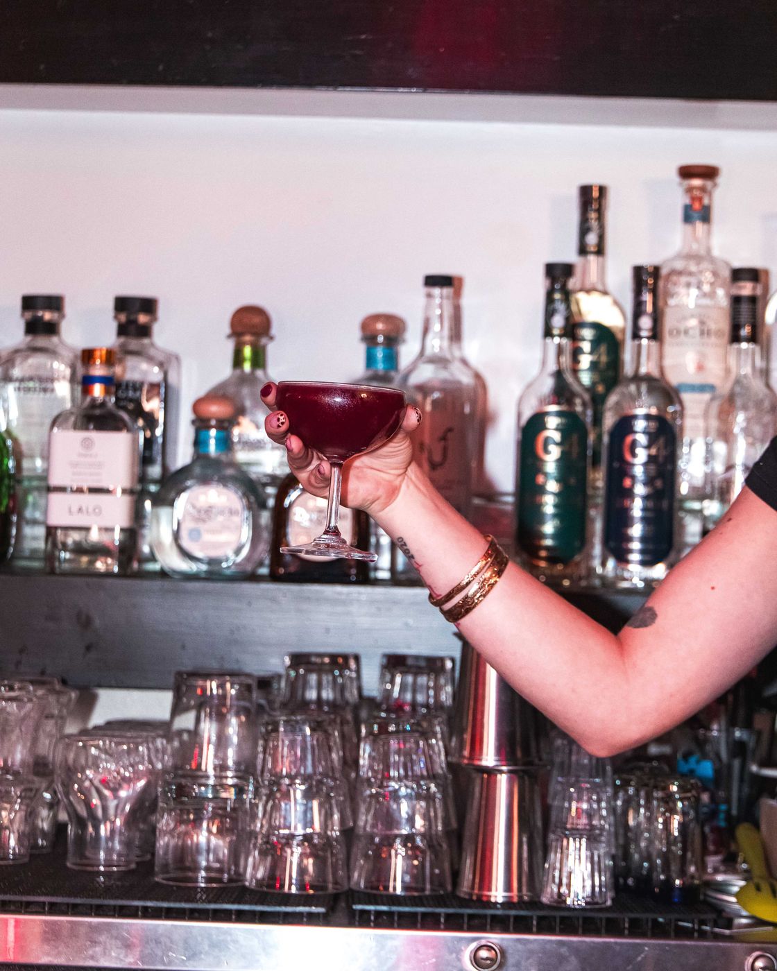 A bartender raising a dark coupe cocktail in front of the back bar